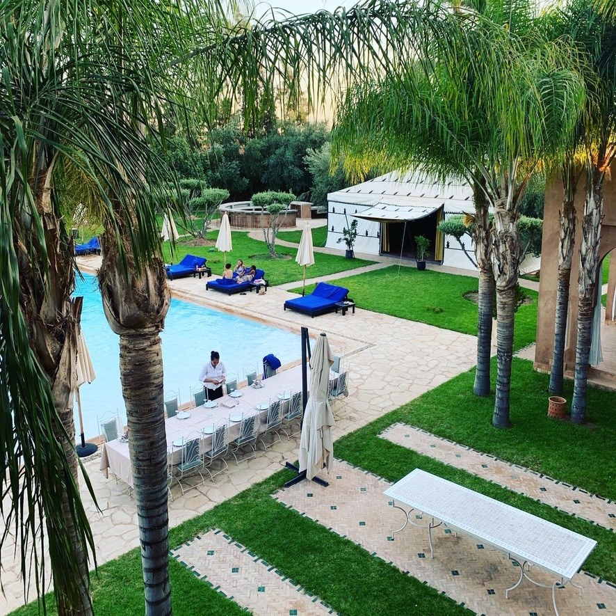 A long table laid with white cloths beside the pool at Palais Tika, under palm trees, with a white tent and blue loungers on the lawn beyond
