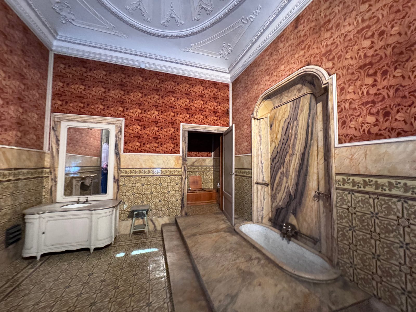 A bathroom at the Villa Reale with a marble bath in an arched alcove of veined marble, patterned red walls and a stucco ceiling