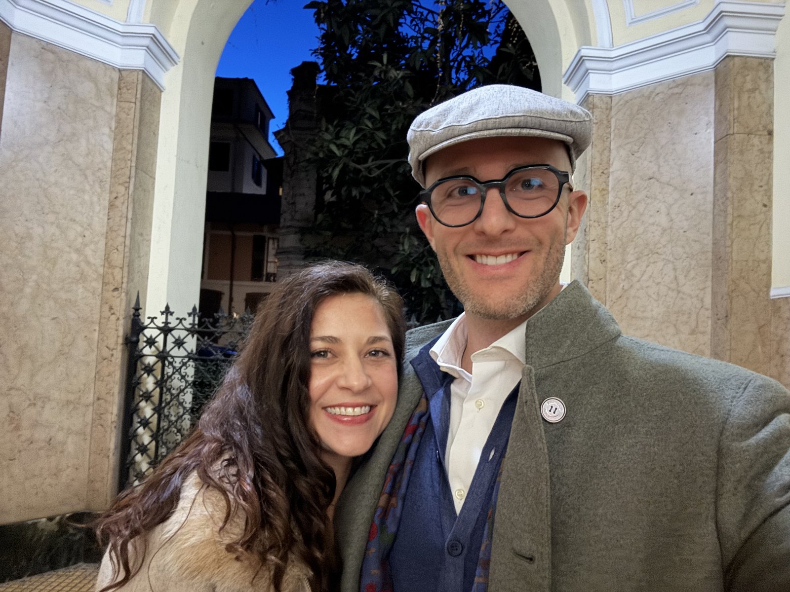 Teri resting her head on Thomas's shoulder under a pale stone arch, Thomas in a flat cap and a green coat